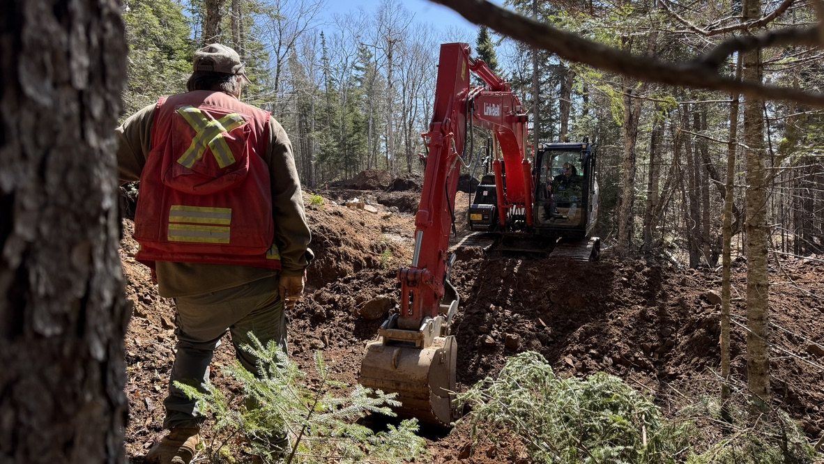 Excavator cutting a trench on an active exploration program while a crew member supervises